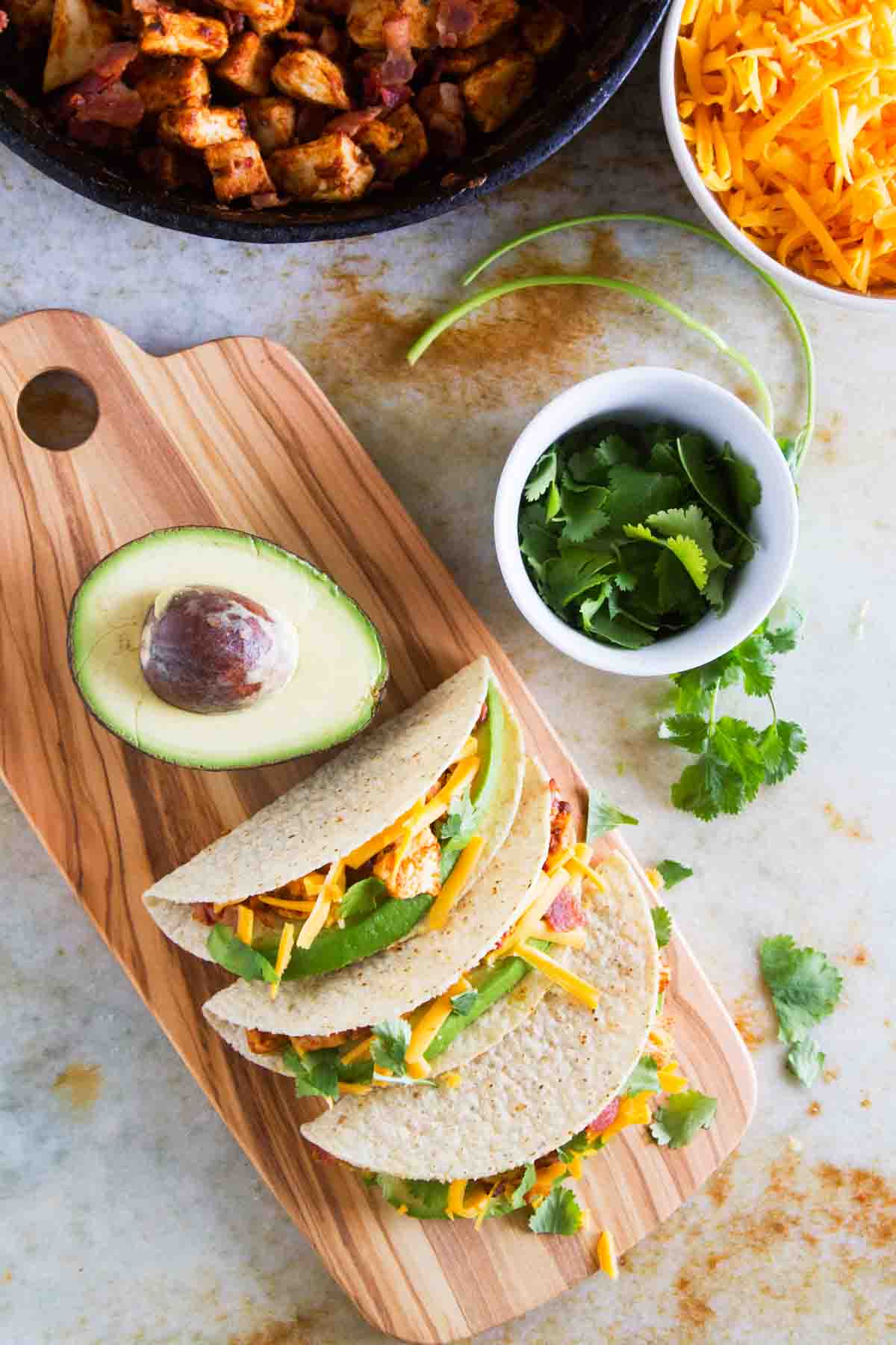 Chicken Bacon Avocado Tacos on a cutting board with avocado, cilantro, and cheese.