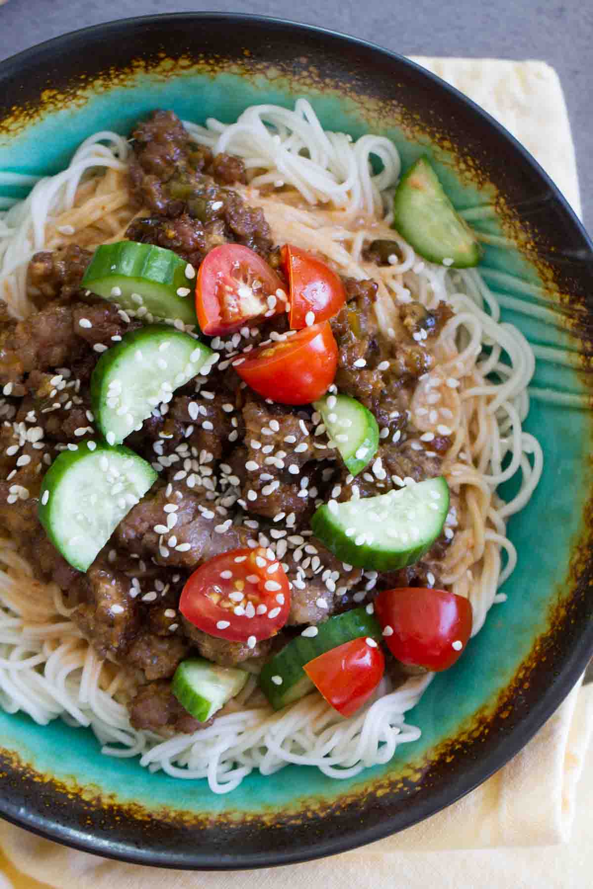 Spicy Pork Noodle Bowl topped with cucumbers, tomatoes, and sesame seeds.