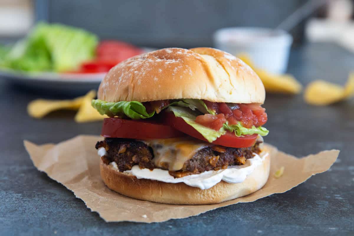 Taco burger with cheese topped with lettuce, tomatoes, salsa, and sour cream.