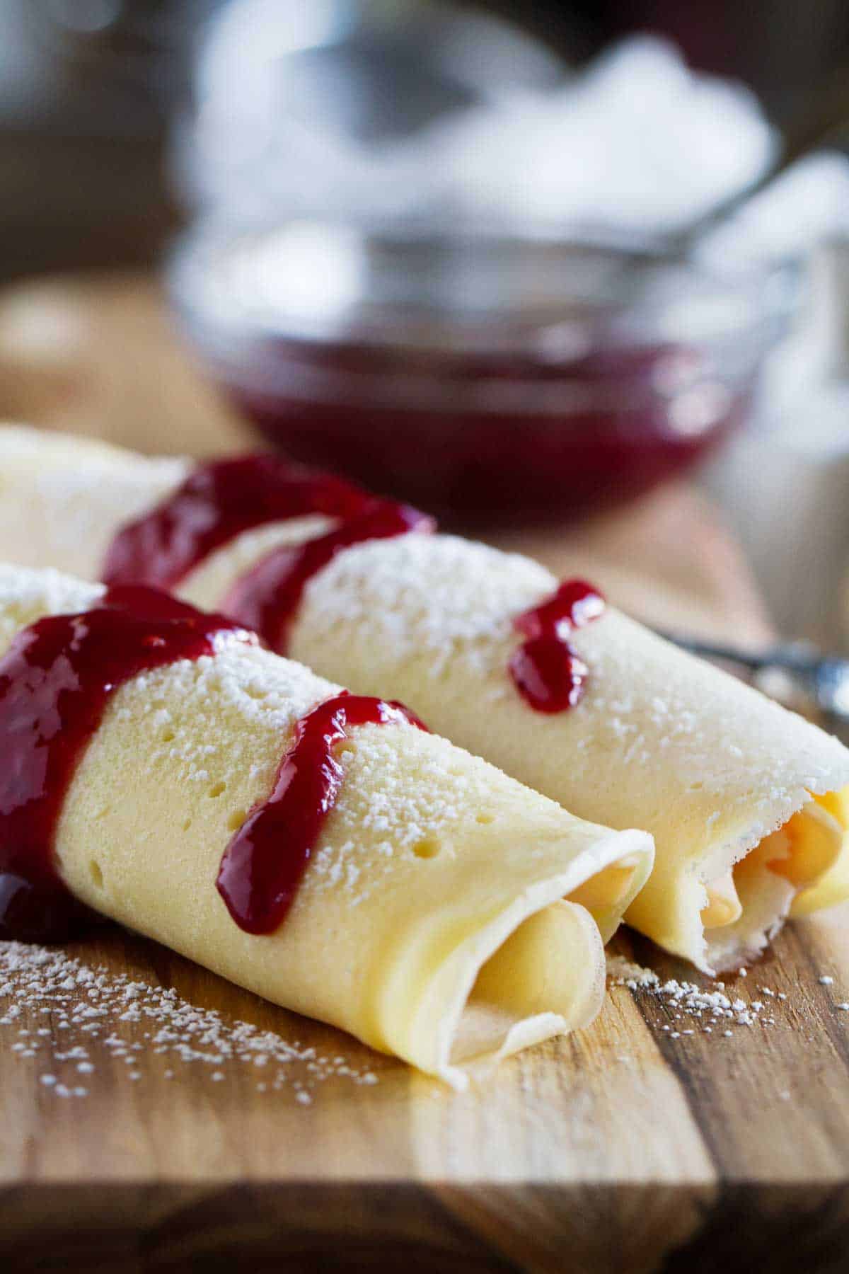 Two rolled monte cristo crepes on a cutting board, topped with jam and powdered sugar.