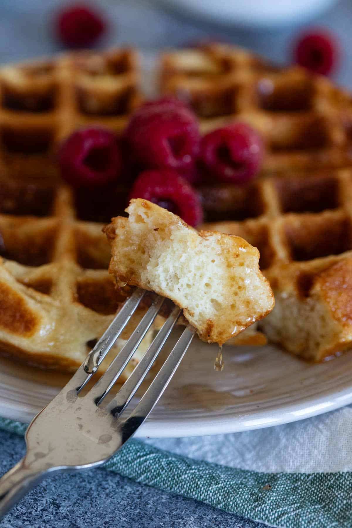 Bite of Belgian waffle on a fork with a drip of syrup coming off of it.