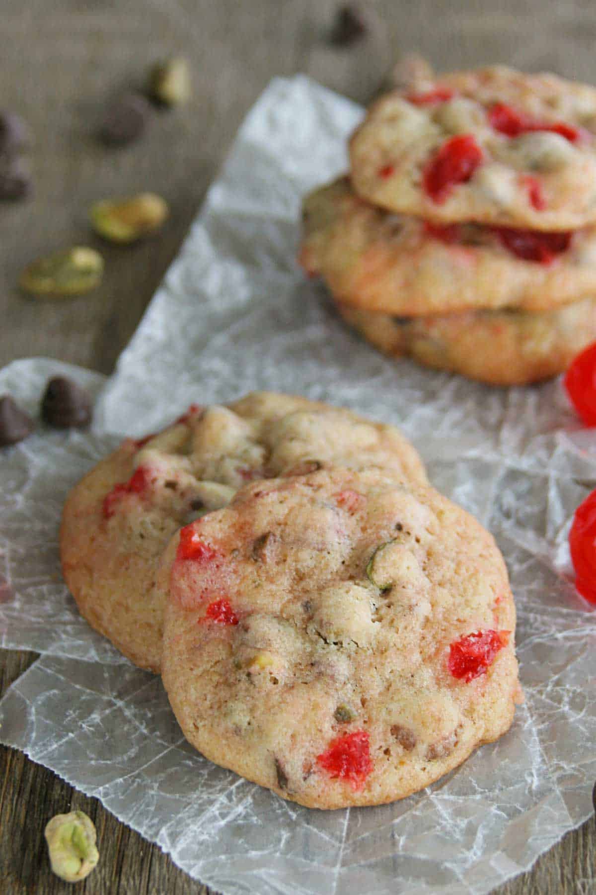 Cherry Chocolate Nut Cookies sitting on each other, with pistachios, cherries, and chocolate chips surrounding them.