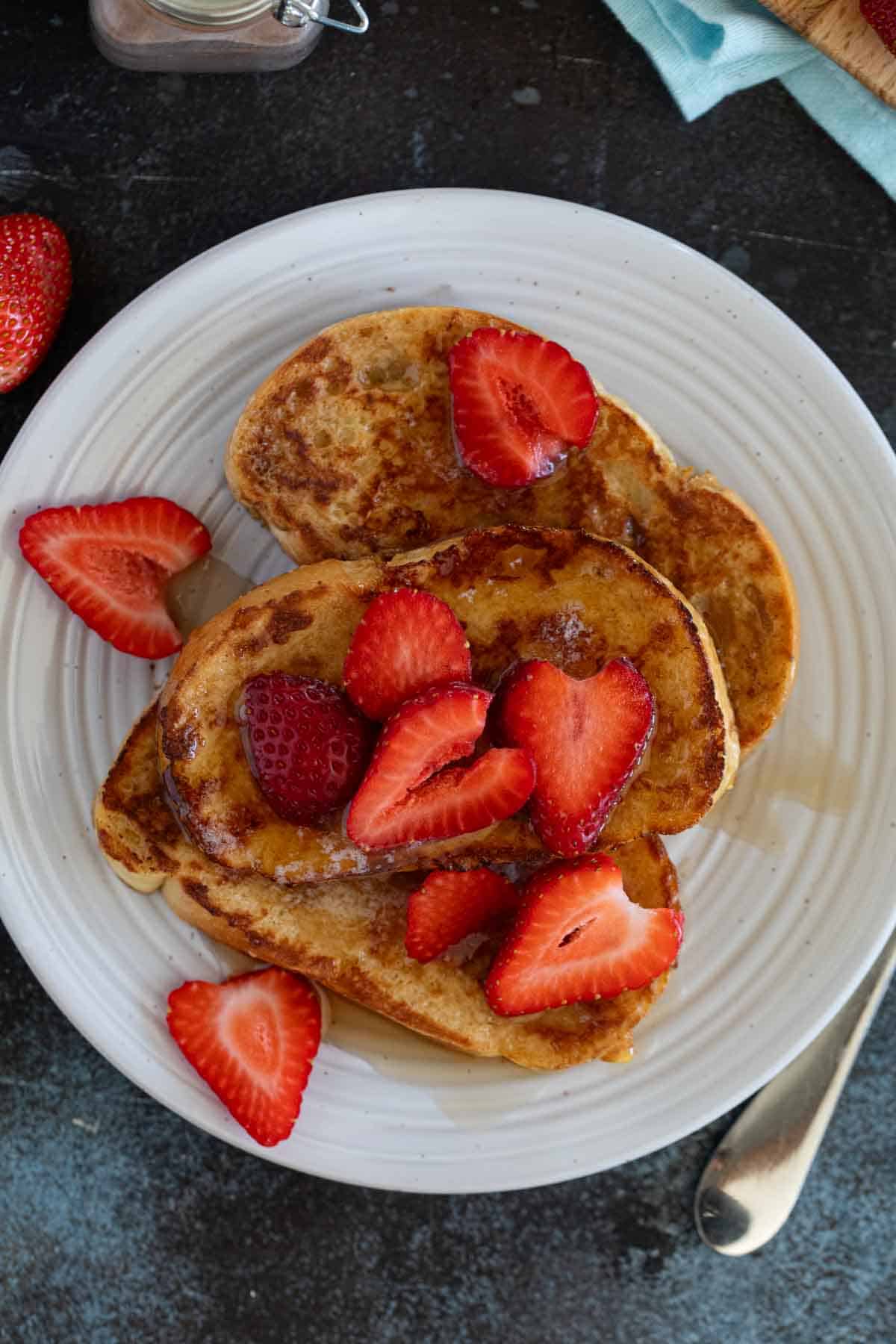 Slices of French Toast on a plate, topped with sliced strawberries and syrup.