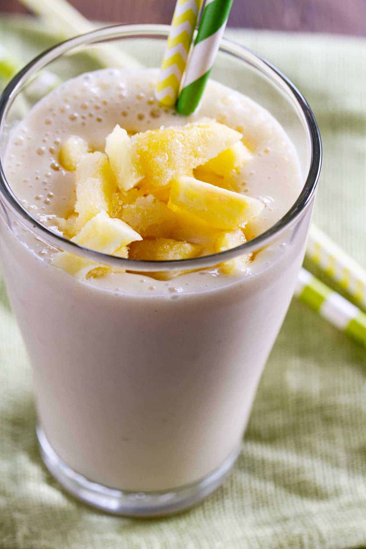 Glass filled with pineapple banana smoothie, topped with more pineapple and with two straws.